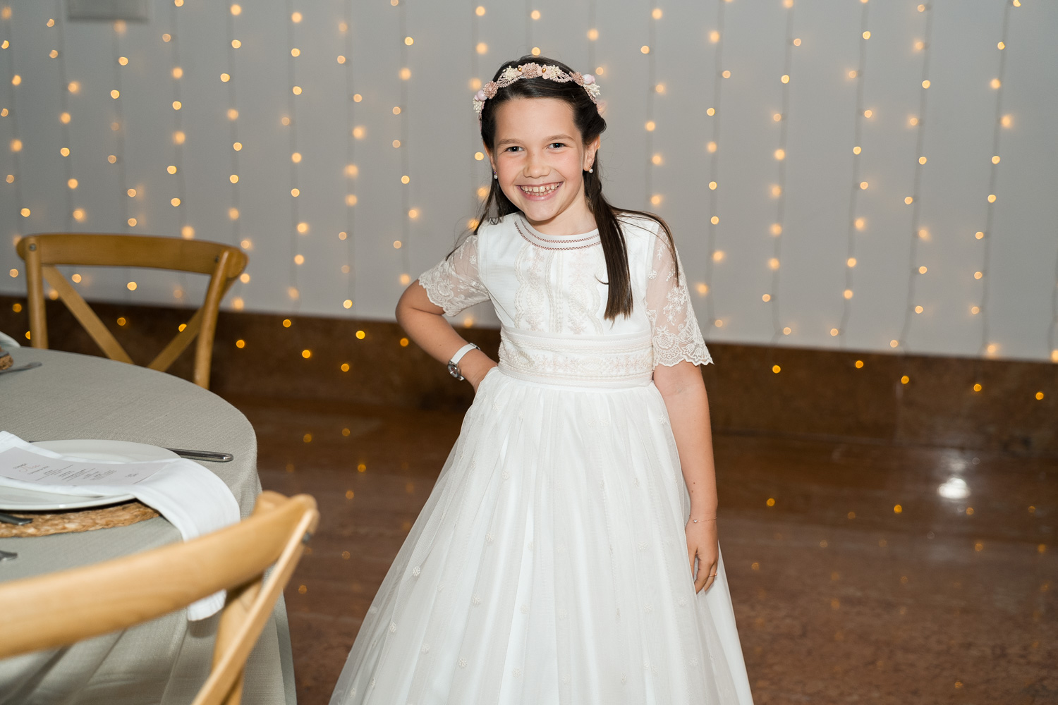 niña posa y ríe con vestido de comunión en salón con luces de fondo